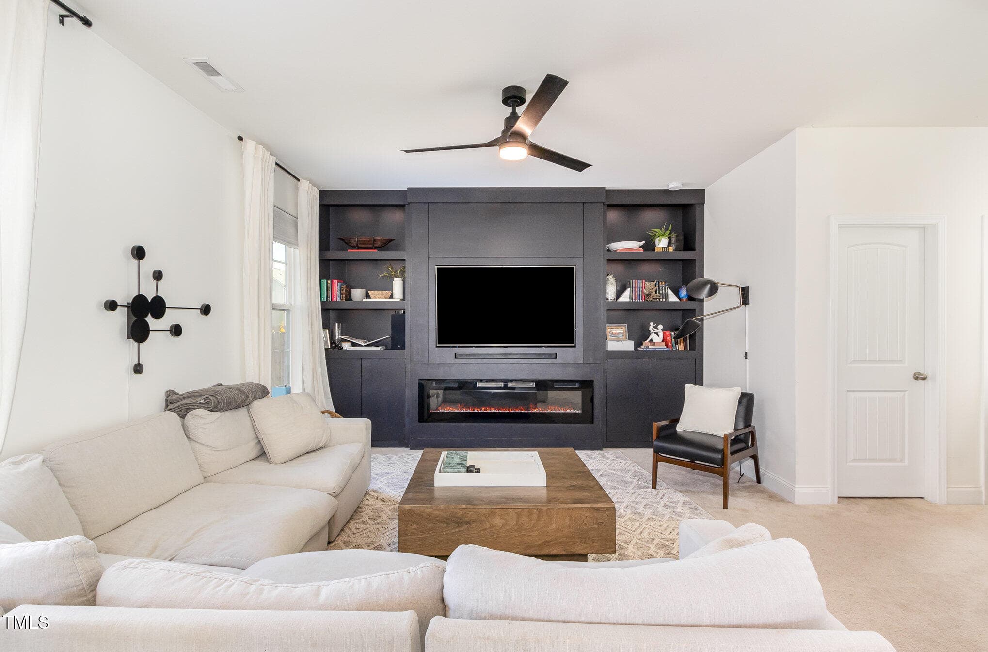 A modern living room with a white sectional sofa, a wooden coffee table, a black accent chair, and a black built-in entertainment center featuring a TV and shelves, with a ceiling fan above.