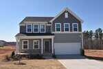 Two-story modern house with gray siding, white trim, and stonework at the base. It has a two-car garage, a small covered front porch, and minimal landscaping. The sky is clear and the lot is newly developed.