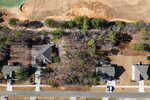 Aerial view of suburban houses with gray roofs, driveways, and lawns; a wooded area separates the homes from a dirt area and golf course in the upper part of the image.
