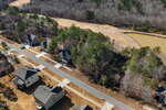 Aerial view of a suburban neighborhood with houses, driveways, and a road. Trees surround the area, and a pond is visible to the right near a cleared field. Some yards appear brown, with scattered cars in driveways.