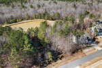 Aerial view of a residential area next to a forested landscape, with a pond partially surrounded by trees and a paved road running alongside several houses.