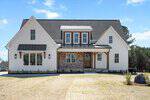 A modern farmhouse-style home with white siding, brick accents, black metal roof awnings, and a covered front porch supported by wooden columns. The house sits on a sparse lawn with minimal landscaping.