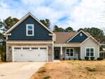 A modern one-story house with dark blue siding, stone accents, and a two-car garage. There is a covered front porch with two white rocking chairs and a lawn with sparse grass. Trees are visible in the background.
