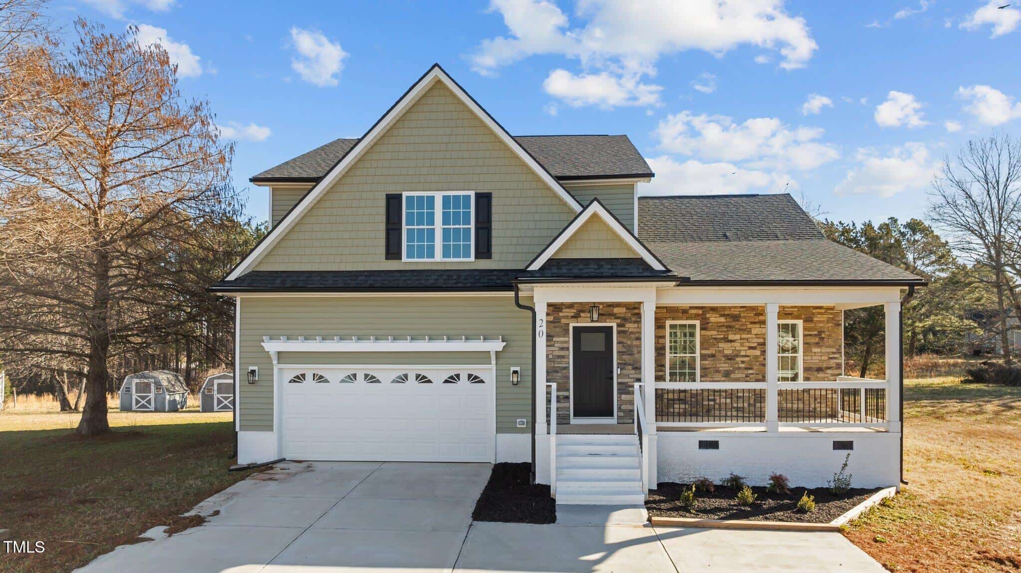 A two-story suburban house with green siding, stone accents, a front porch, white garage door, and a concrete driveway. The yard has sparse landscaping and two sheds are visible in the background.