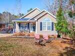 A single-story beige house with white trim and dark blue shutters features a covered porch with chairs and a wreath on the door. In the yard, two red lawn chairs face a metal fire pit. Leafless trees and some greenery surround the home.