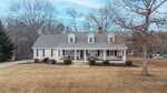 A beige, one-and-a-half-story suburban house with dormer windows, a covered front porch, and a shingled roof. The yard is mostly brown grass with sparse shrubs and trees, and a driveway runs along the left side.