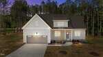 A two-story, light gray house with a dark roof, double garage, and front porch is set on a large lot with minimal landscaping, surrounded by tall pine trees at sunset.