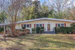 A single-story house with light yellow siding, red shutters, a covered carport on the left, front steps leading to a wooden door, and mature trees and shrubs in the front yard.