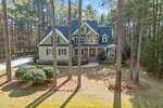 A two-story house with a front porch, gabled roof, and stone accents sits among tall pine trees. There is a curved driveway and landscaped bushes in the front yard, with pine needles covering the ground.