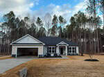 A single-story house with a dark roof and light-colored siding sits in front of a driveway and lawn, with pine trees and a cloudy sky in the background. There is a two-car garage and a covered entrance.