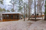 A small cottage with a screened porch sits on a gravel lot surrounded by trees, next to a travel trailer on a wooden deck. The area is quiet and wooded, with leafless trees and overcast skies.