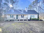 A white, one-and-a-half story house with a covered front porch, green door, dormer windows, and a shingled roof, surrounded by trees and a patchy lawn under a clear sky.