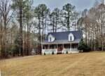 A two-story house with a covered front porch and dormer windows sits on a spacious lawn surrounded by tall trees on a cloudy day. A driveway leads up to the house.