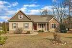 Single-story house with tan siding, stone accents, and brown shutters, located on a spacious, partially landscaped yard with a concrete walkway leading to the front door and rocking chair on the porch. Trees border the property.