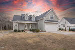 Two-story gray house with white trim, double garage, dormer windows, and a covered front porch. The front yard is mostly bare with some shrubs. The sky is colorful with pink and orange clouds at sunset.