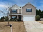 A two-story suburban house with beige siding, a covered front porch, black shutters, a white double garage door, a bare tree in the front yard, and a driveway leading to the street.