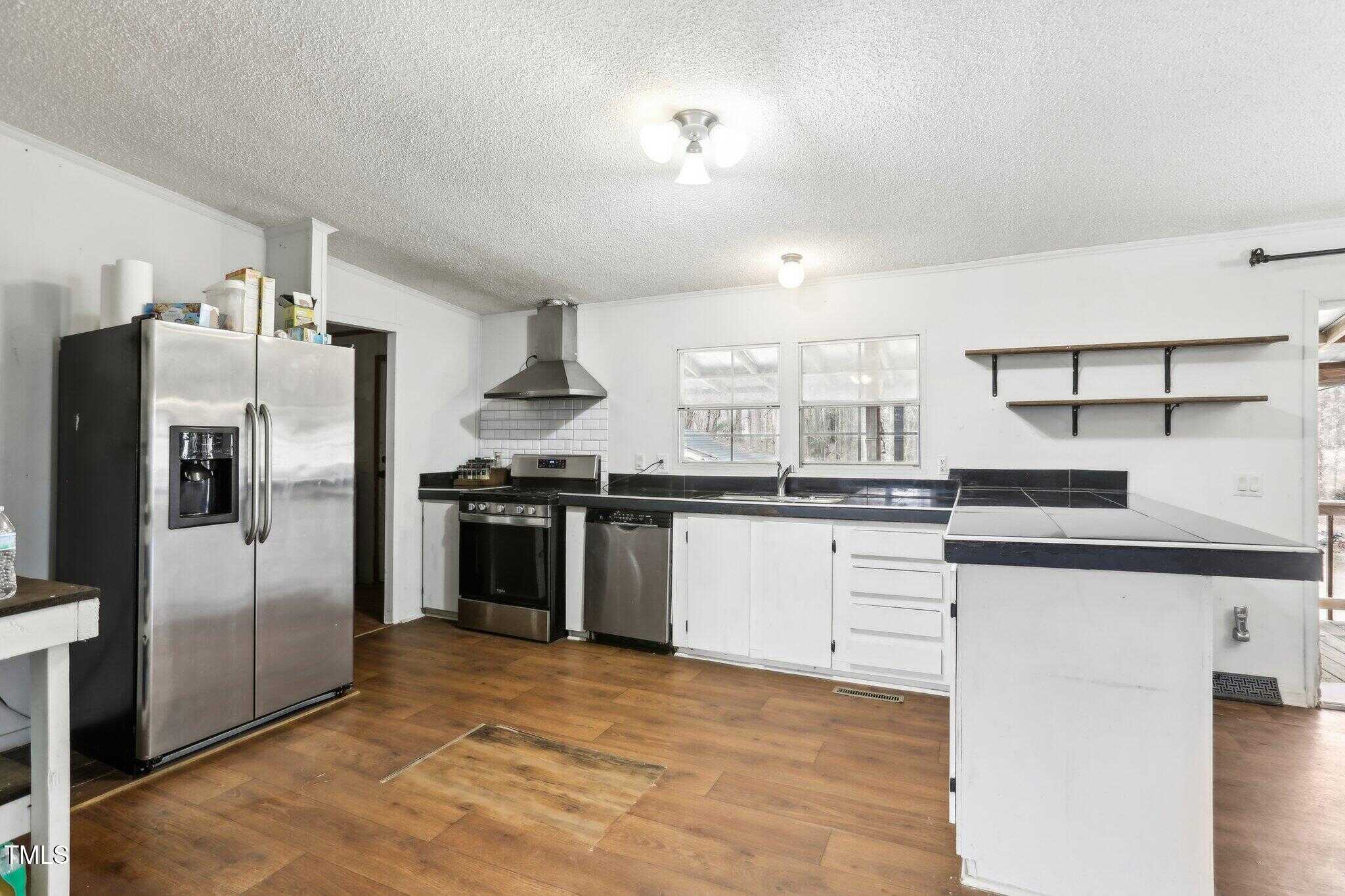 A kitchen with stainless steel refrigerator, stove, and range hood, white cabinets, open shelves, black countertops, wood flooring, and a ceiling light fixture. Two windows provide natural light above the sink area.