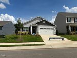 Single-story house with gray siding, white trim, and a black accent above the garage. It has a covered front porch, neatly landscaped lawn, and a driveway leading to a double garage. A promotional sign is on the front yard.