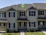 A row of two-story tan townhouses with dark shutters and small front porches; a green location marker with a house icon is pointing to the center unit on the upper floor.