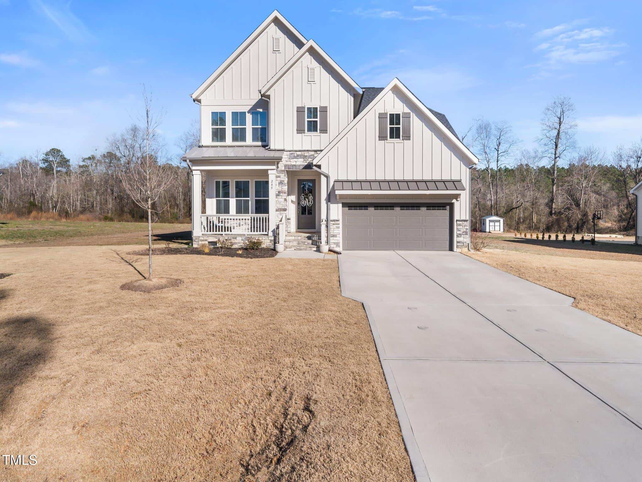 A two-story beige house with white trim, a front porch, double garage, and a large concrete driveway. The yard is covered with dry grass, and trees are visible in the background under a clear blue sky.