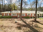 A single-story brick house with white trim sits among tall trees. The yard is mostly bare soil with scattered leaves and some greenery near the house. A carport with a vehicle is attached on the right side.