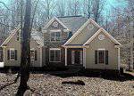 A two-story house with light beige siding, stone accents, white trim, and a dark shingle roof, situated in a wooded area with leafless trees and a covering of dry leaves on the ground.