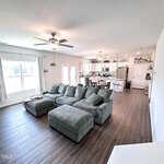 Open-concept living area with a large green sectional sofa and ottoman on wood flooring, adjacent to a kitchen with white cabinets and stainless steel appliances, and a dining table; ceiling fan above.