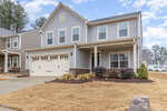 A two-story suburban house with gray siding, white trim, and a two-car garage. The front yard has dry grass, a small shrub garden, and a covered porch. Other houses and trees are visible in the background.