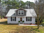 A white, one-and-a-half-story house with a gray metal roof and dormer window, two front windows with black shutters, two small potted plants by the entrance, and a leafless tree in the front yard. Pine trees are in the background.