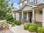 A two-story beige house with large windows, stone accents, and a covered front porch. The yard has neatly trimmed bushes, small trees, and a paved walkway with white gravel landscaping.