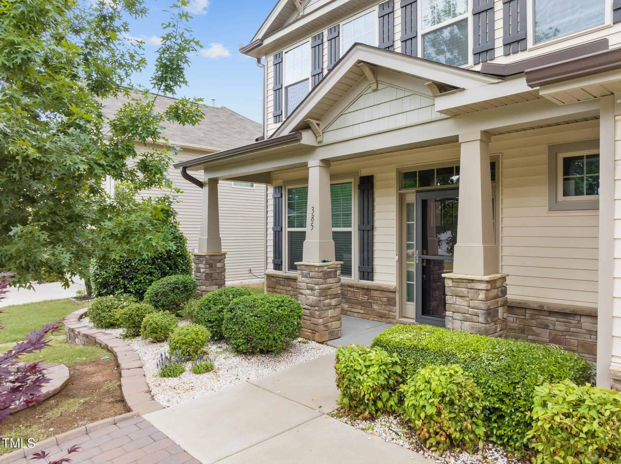 A two-story beige house with large windows, stone accents, and a covered front porch. The yard has neatly trimmed bushes, small trees, and a paved walkway with white gravel landscaping.