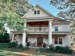 A two-story white house with large columns, a covered front porch, an upper balcony, and a dark wooden front door. The yard has green plants and trees, and there is a ceiling fan on the porch.