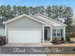 Single-story house with gray siding, white trim, and a two-car garage. Small covered front porch with columns, landscaped yard, and a background of tall pine trees. Banner text reads, "Ranch - Shows Like New!.