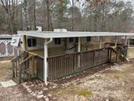 A white travel trailer is parked under a metal carport with a wooden wheelchair-accessible ramp and railing. The trailer is located in a wooded area, with a shed visible in the background.