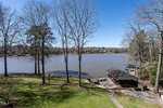 A lakeside view with leafless trees in the foreground, a grassy yard, and a small dock and covered boathouse on the right. Calm water and distant wooded shoreline are visible under a clear blue sky.