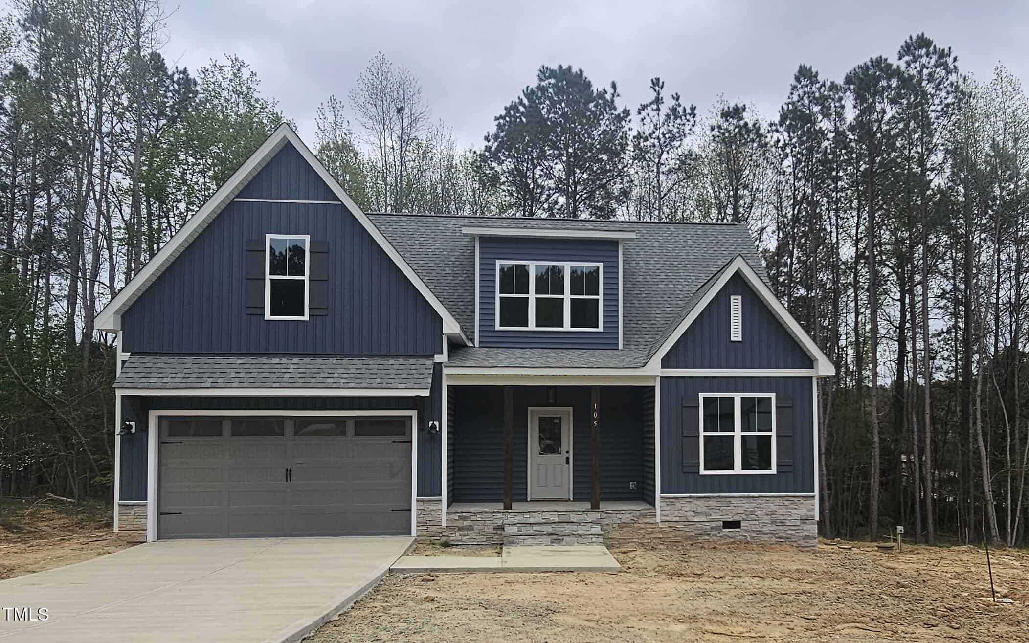 A two-story blue house with white trim, a gray shingle roof, an attached two-car garage, and a concrete driveway. It is surrounded by bare ground and trees in the background.