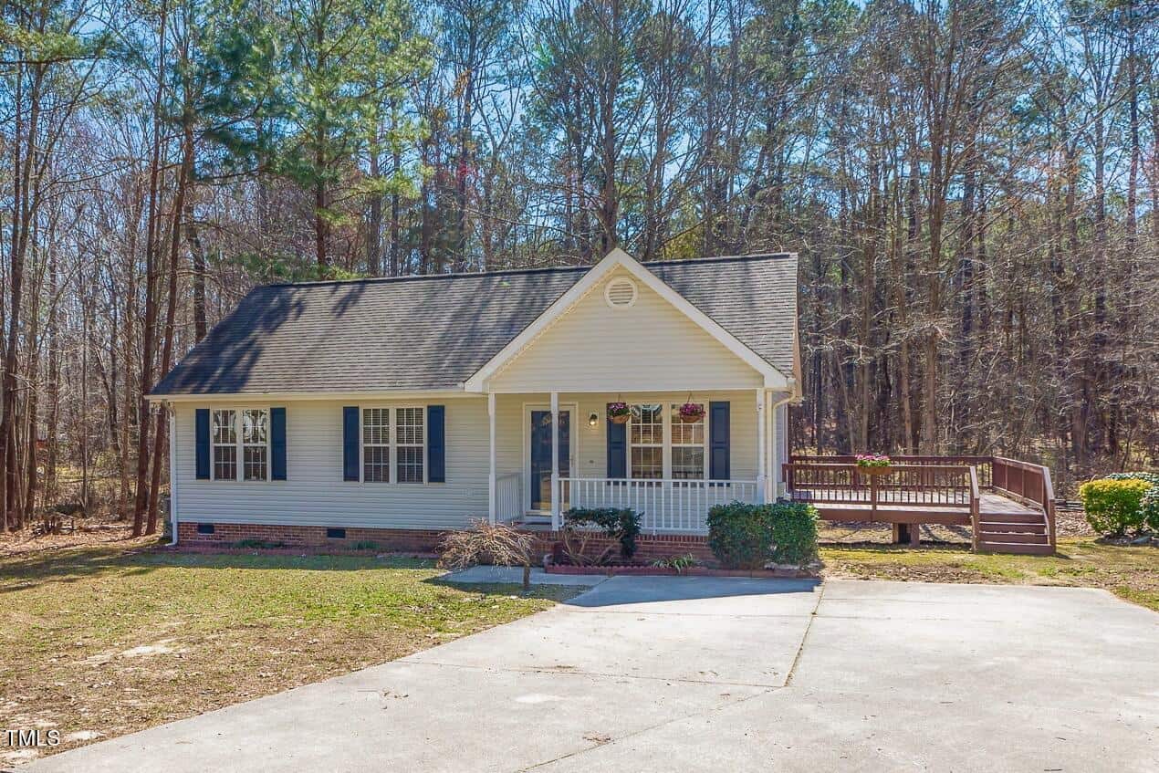 A small, single-story house with white siding, black shutters, and a front porch. A wooden ramp leads to the side of the house. The home is surrounded by trees and a large concrete driveway.