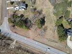 Aerial view of a vacant, partially wooded lot outlined in red at the corner of two roads, with a house and driveway on the adjacent lot; trees and open grass are visible within the outlined area.