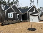 A modern single-story house with dark vertical siding, stone accents, white trim, and a two-car garage. The yard is covered in straw with a small tree planted near the driveway. Tall pine trees are in the background.