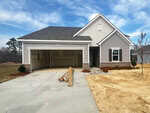 A newly constructed, single-story house with gray siding, a two-car garage with doors open, and an unfinished yard with patches of bare soil and a small tree in front. Construction materials lie on the driveway.