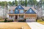 Two-story suburban house with beige siding, blue accents, and stone facade. Features a covered front porch, double garage, and a well-maintained lawn. Tall trees line the background.