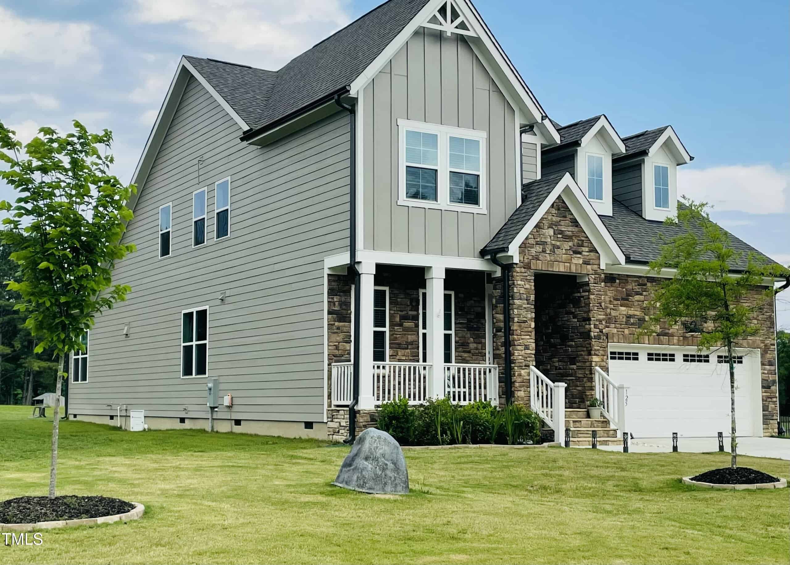 A two-story modern suburban house with gray siding and stone accents, white trim, and a covered front porch. The yard is neatly mowed with small trees and a large decorative rock on the lawn.