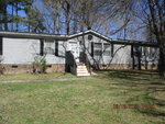A single-story manufactured home with gray siding and a brick foundation. The front entrance has a small porch with steps. The yard is grassy and surrounded by tall trees. The photo is dated March 18, 2025.