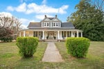 A large two-story house with white siding, a wraparound front porch, and dark roof shingles. The porch has columns and rocking chairs. A concrete walkway leads to the front steps, bordered by trimmed bushes and a grassy yard.