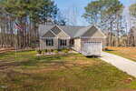 Single-story house with beige siding, gray roof, a covered front porch, and an attached two-car garage. A concrete driveway and ramp lead to the entrance. An American flag is on a flagpole in the grassy front yard, surrounded by tall trees.