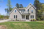 A modern two-story house with gray siding and white trim sits on a green lawn with minimal landscaping, surrounded by tall trees under a clear blue sky.