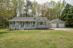 Single-story gray house with white trim and front porch, surrounded by grass and trees, with a detached two-car garage on the right side under a partly cloudy sky.
