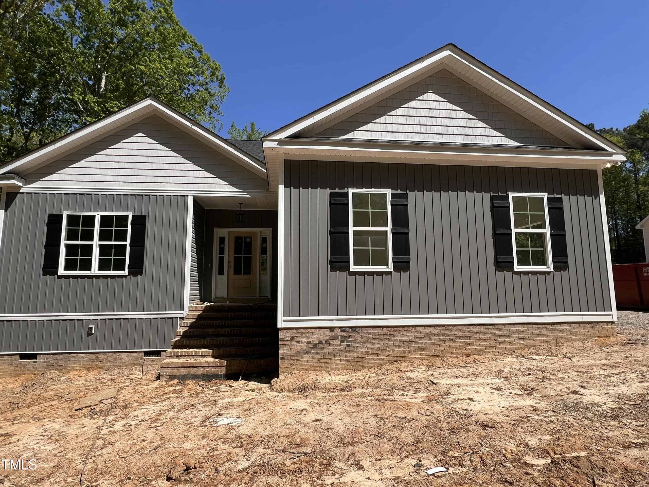 A single-story gray house with white trim and black shutters, featuring a gabled roof, brick foundation, and front steps leading to a white double-door entry. The yard is bare dirt, and trees are visible in the background.