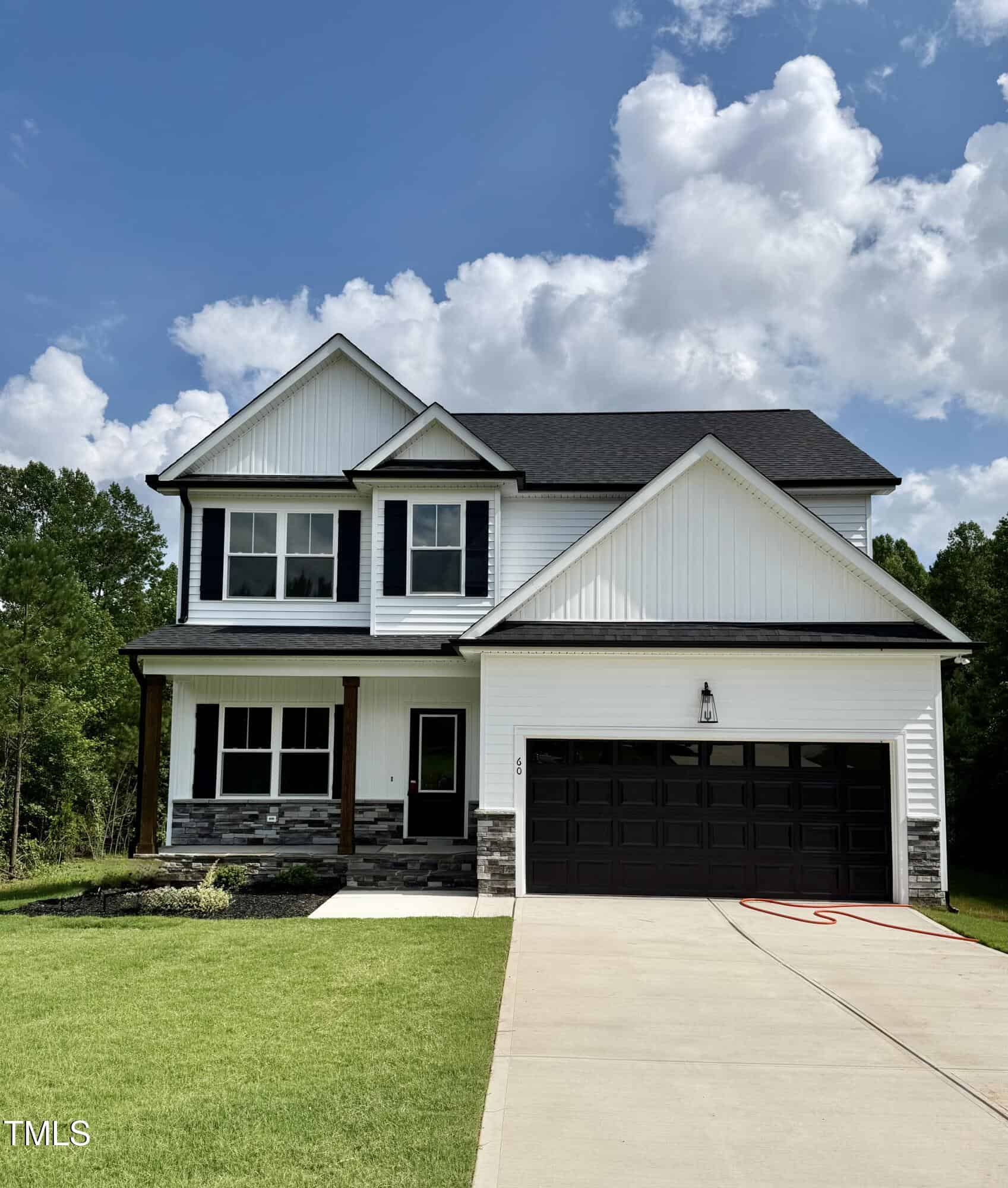 A two-story modern suburban house with white siding, black trim, and a dark garage door. The home has a covered front porch, a manicured lawn, and a concrete driveway. Trees and blue sky with clouds are in the background.
