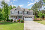 Two-story suburban house with gray siding, stone accents, and a white two-car garage. The home has a front porch, green lawn, flower beds, and is surrounded by trees and a wooden fence.
